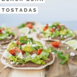 Three black bean tostadas on a wooden board with tomatoes and avocados