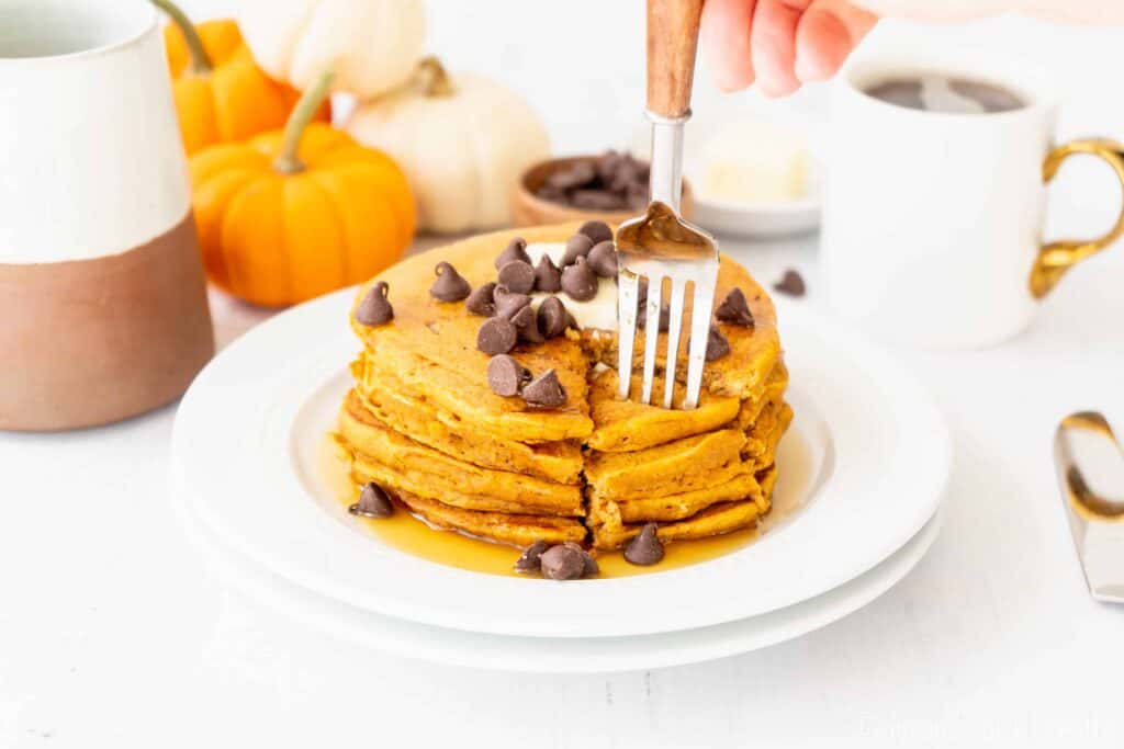 a fork taking a bite of pumpkin pancakes on a white plate with a cup of coffee