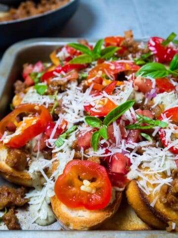 Take game day to another level with these delicious Italian Nachos with Spicy Sausage and Pesto Alfredo. They're everything you love about nachos, but they'd make any Nonna proud! Toasted bread is topped with spicy Italian sausage, tomato and cherry pepper relish and pesto alfredo! Make a big pan and watch them disappear! | Sip and Spice #gameday #nachos #Italian #pestoalfredo #snacks #dinner