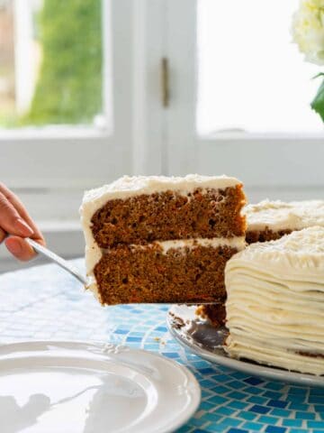 Slice of carrot cake with cream cheese frosting being removed from whole cake on blue table