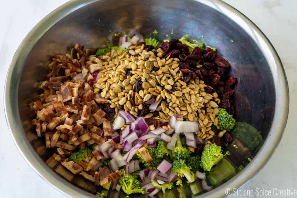 ingredients in a silver mixing bowl - broccoli, sunflower seeds, red onion, bacon, craisins