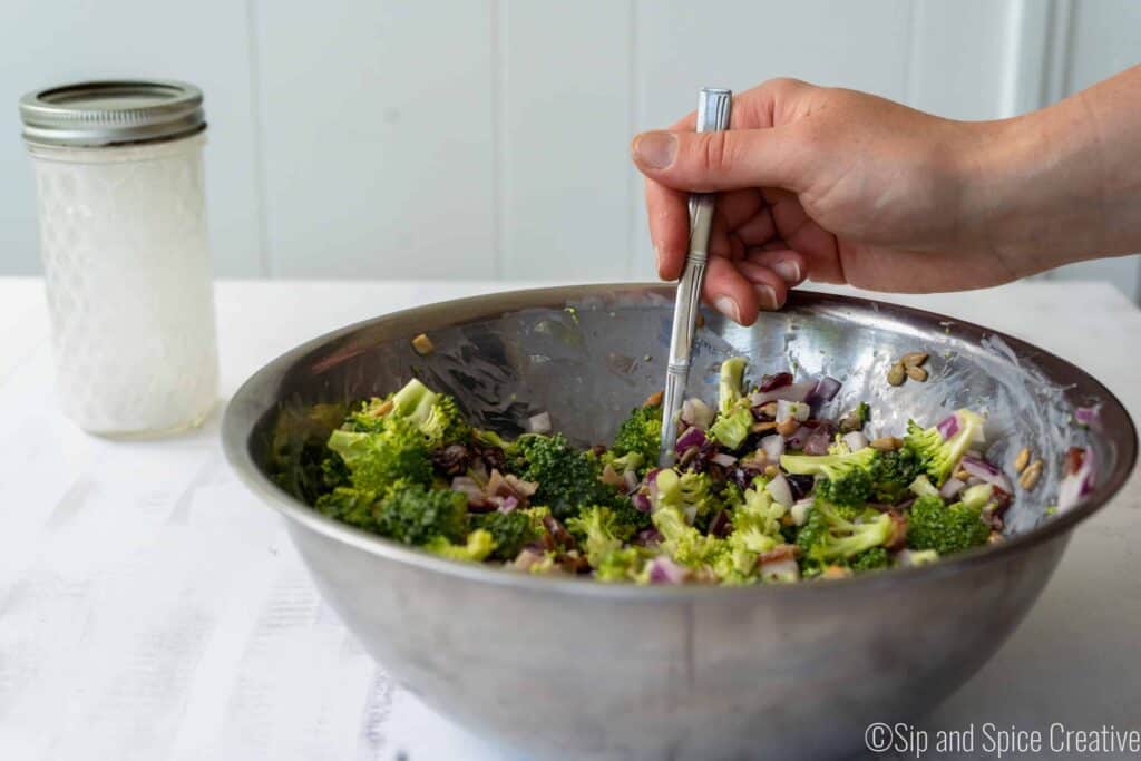 mixing greek yogurt dressing into broccoli salad in a silver mixing bowl