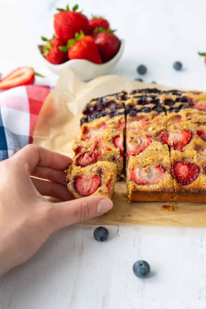 hand grabbing corner slice of snack cake topped with strawberries and blueberries