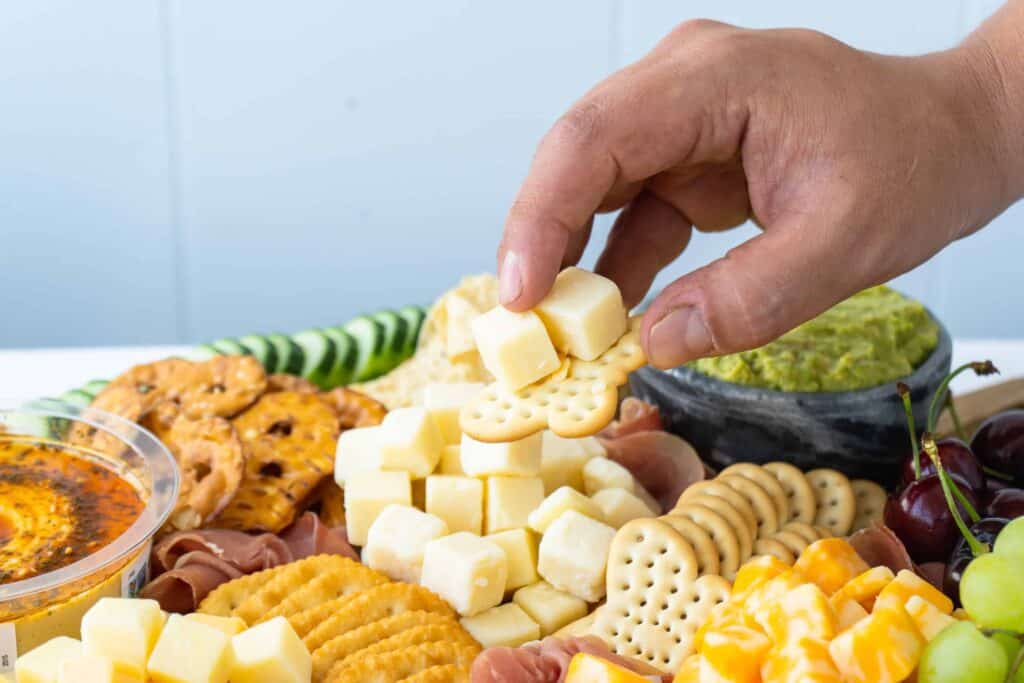 male hand grabbing white cheese cubes and a butterfly cracker over a cheeseboard