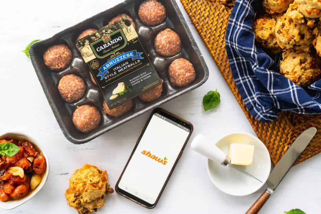 carando meatball packaging next to a bowl of roasted tomatoes, the shaw's app on a phone and a basket of biscuits