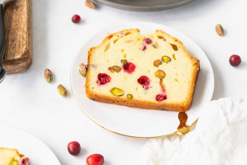 slice of Cranberry Pistachio Pound Cake on a white plate with pistachio and cranberries on the table
