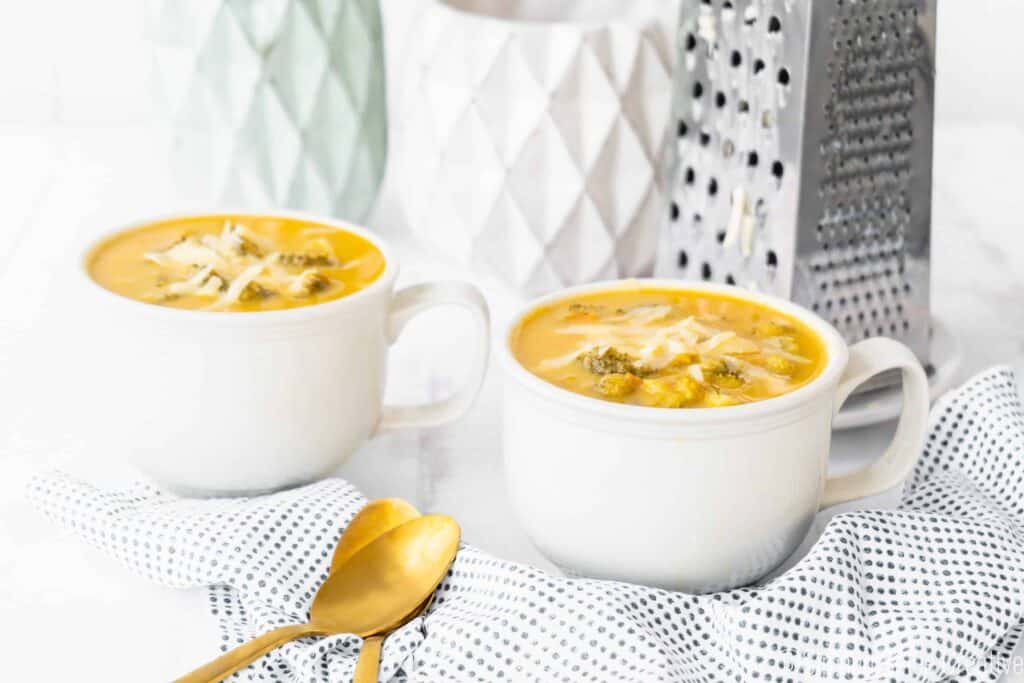 two mugs of broccoli cheddar soup with cheddar cheese and gold spoons