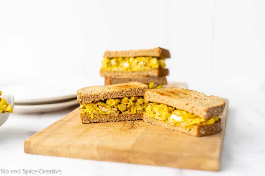 two curried chicken salad sandwiches on wheat bread next to each other on a wooden cutting board