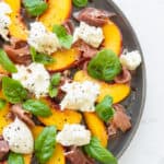 top down view of peach salad with burrata prosciutto and basil leaves on a gray plate