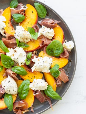 top down view of peach salad with burrata prosciutto and basil leaves on a gray plate