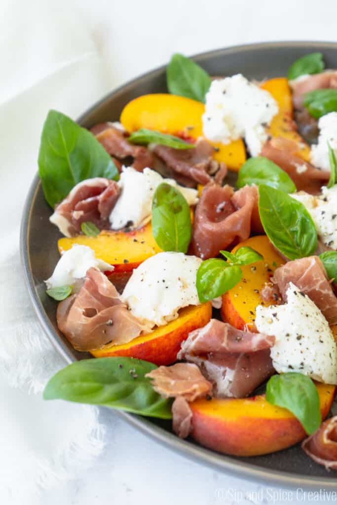 side view of sliced peaches, burrata, prosciutto and basil on a grey plate