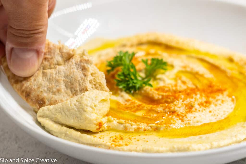 hand scooping hummus from a white bowl with warm pita bread