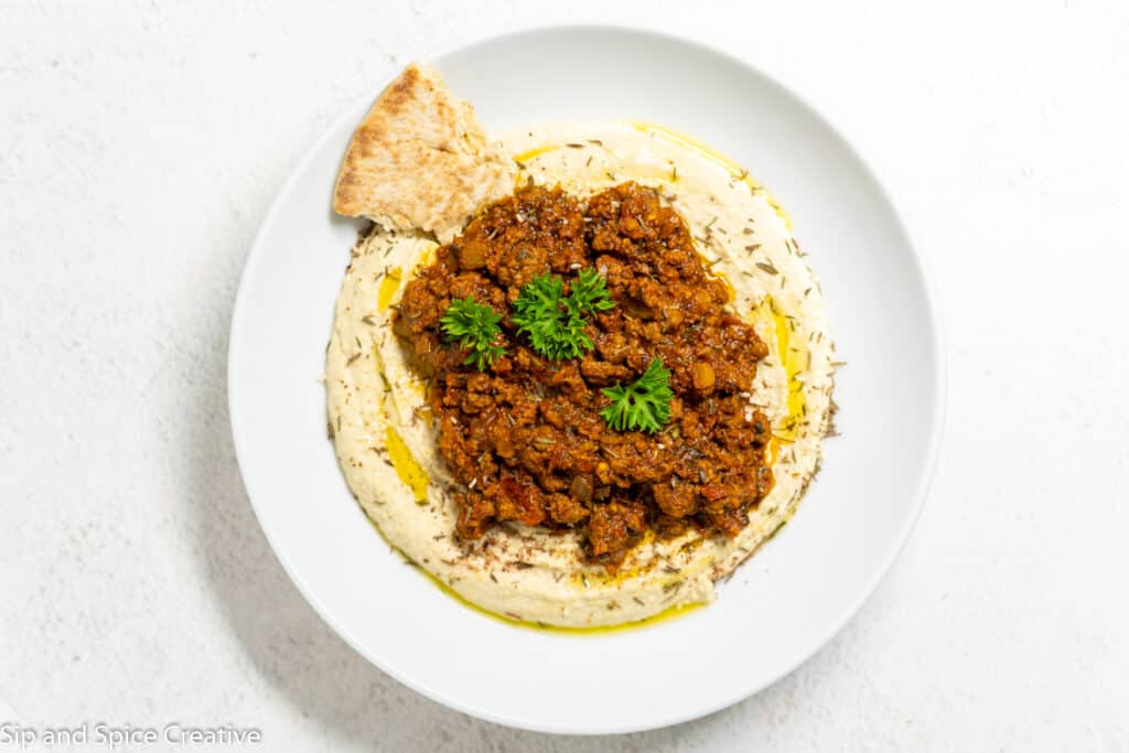 top down view of a white bowl filled with hummus and spicy beef on top with a pita on the side