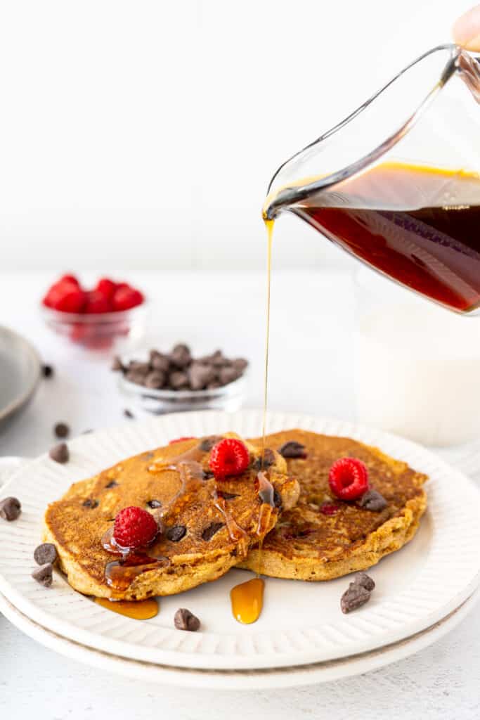 maple syrup pouring from a glass container onto a plate of protein pancakes with raspberries and chocolate chips