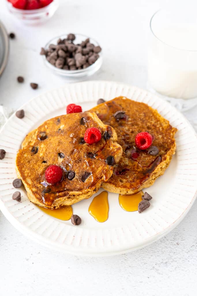 closeup of two protein pancakes on a white plate with raspberries and chocolate chips with a glass of milk