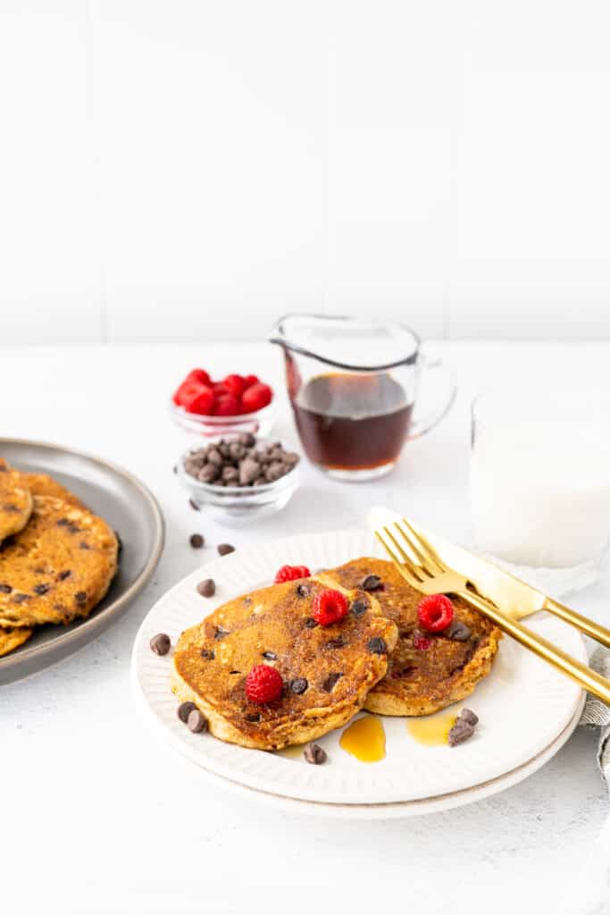 plate of protein pancakes with raspberries and chocolate chips with maple syrup and a gold fork and knife