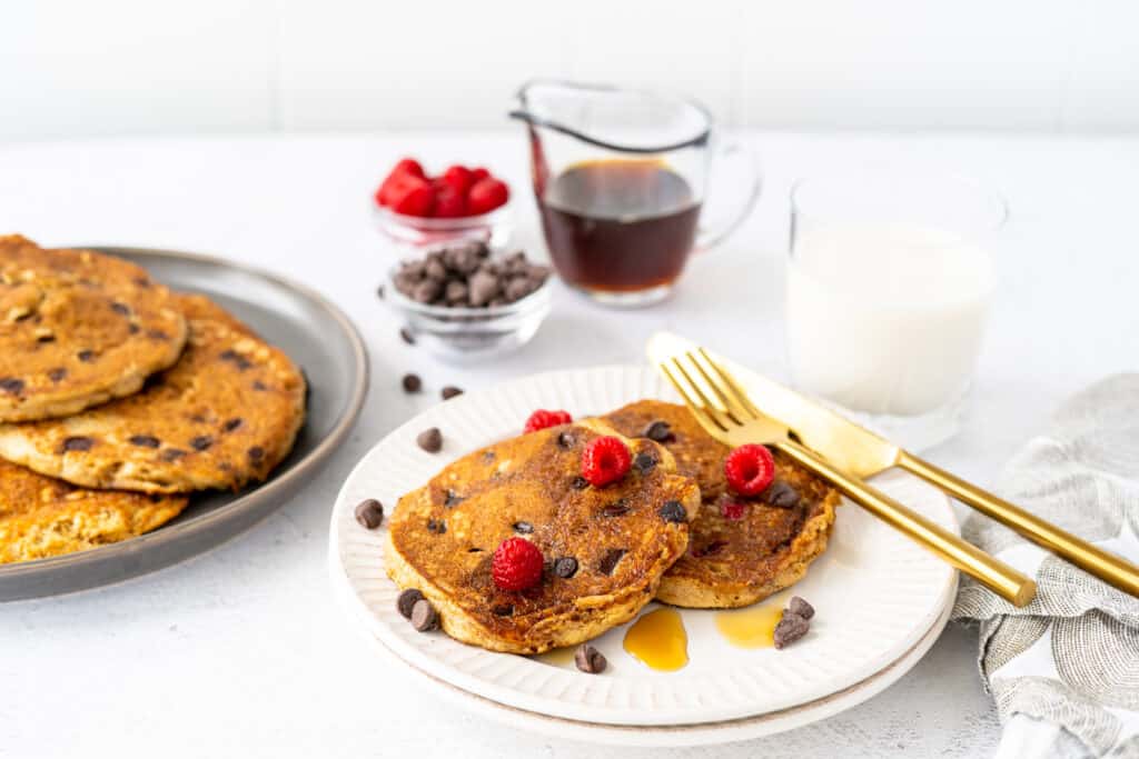 a white plate of protein pancakes with raspberries and chocolate chips with gold silverware and a serving plate full of pancakes behind