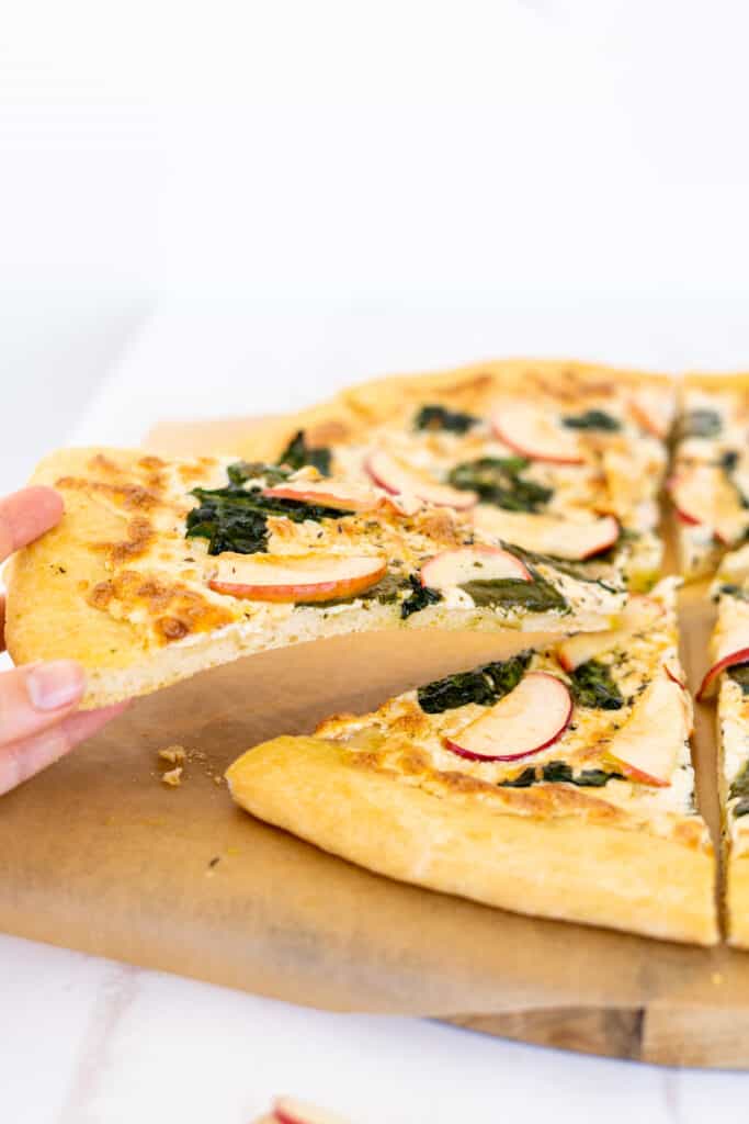 A hand lifting up a slice of pizza topped with spinach and apple on a wooden board