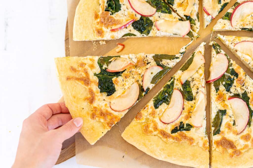 Sliced pizza topped with spinach and apple on a wooden board with a hand pulling out a slice