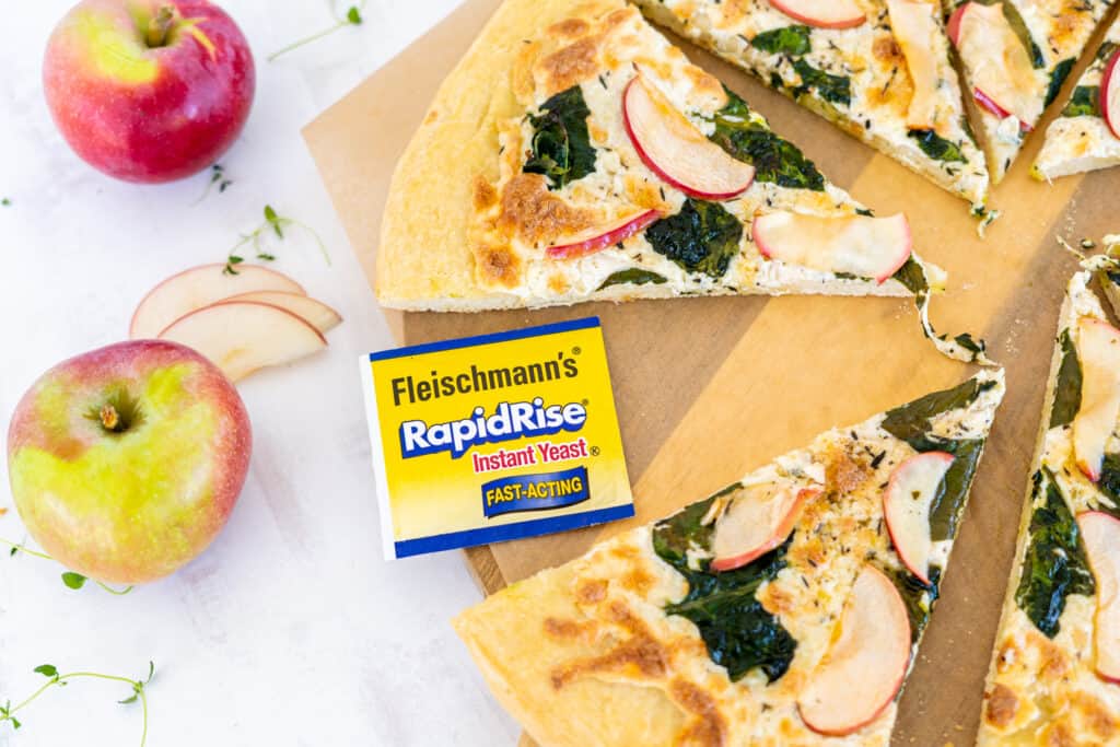 Sliced pizza topped with spinach and apple on a wooden board with a yeast packet surrounded by apples and thyme