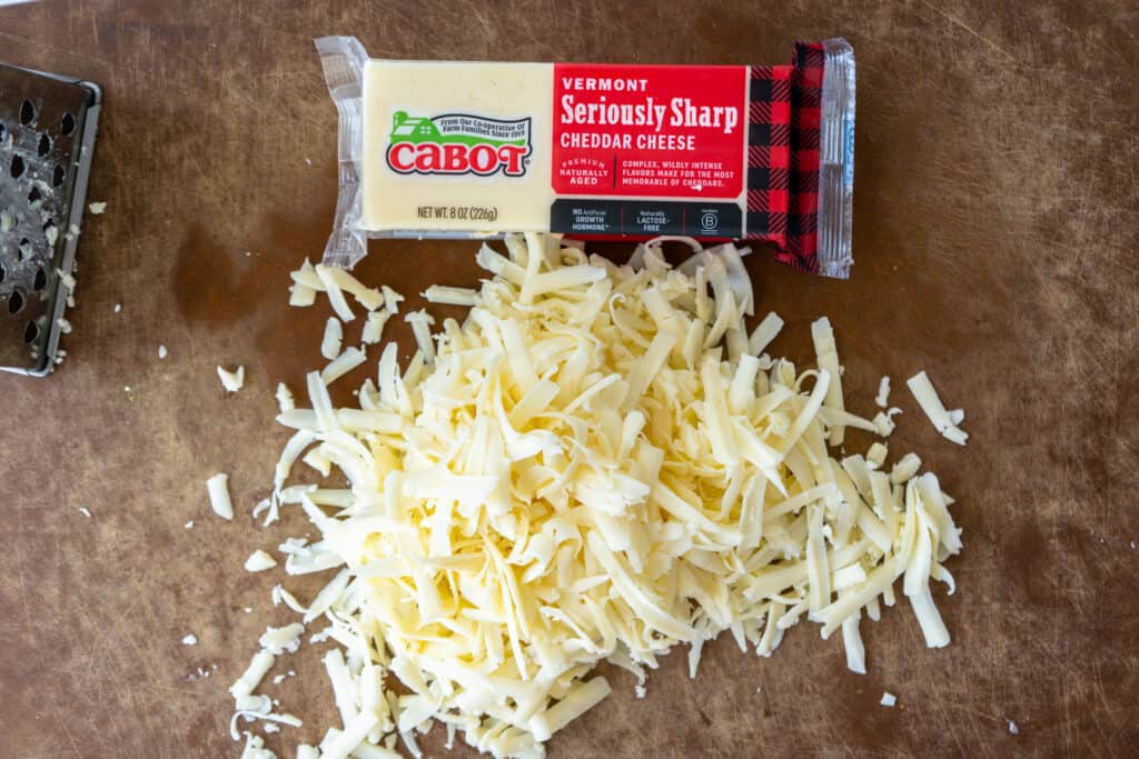 freshly shredded white cheddar cheese on a wooden cuttin board with cabot cheese blocks