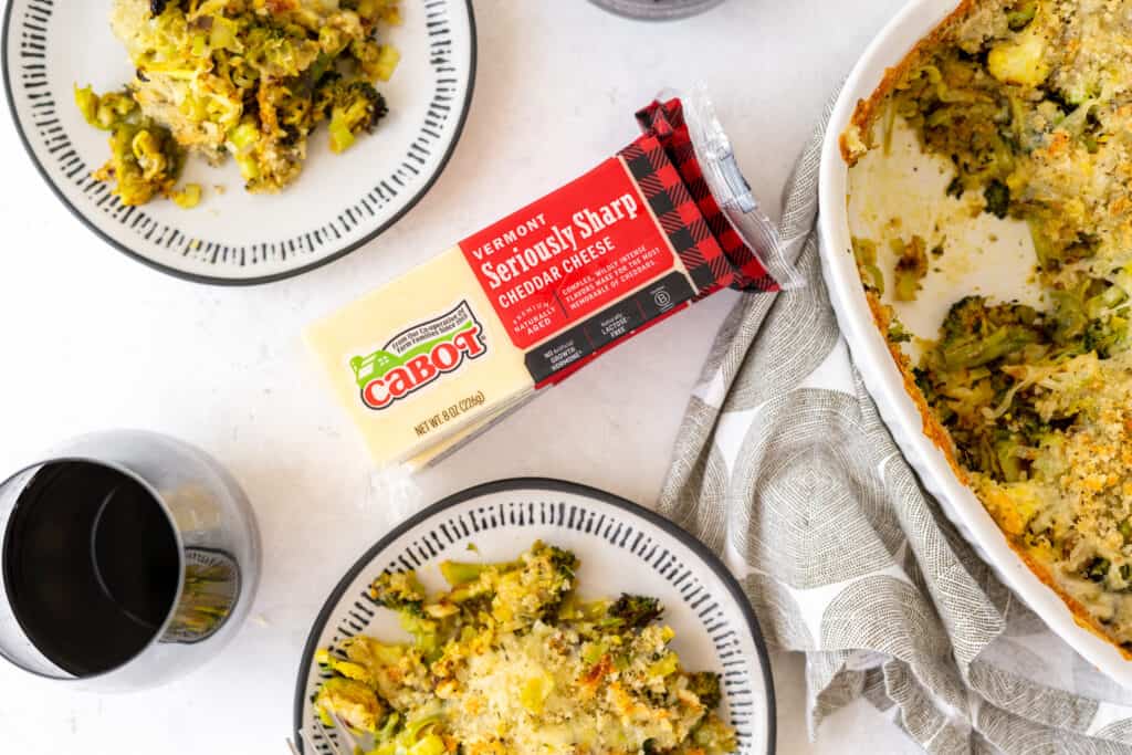 top down view of white tabletop with plates of veggie gratin next to a casserole dish of gratin with cabot cheese blocks in the middle of table
