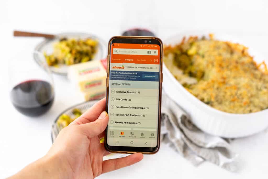 shaw's mobile app on phone being held by a hand in front of a white tabletop with vegetable gratin plated