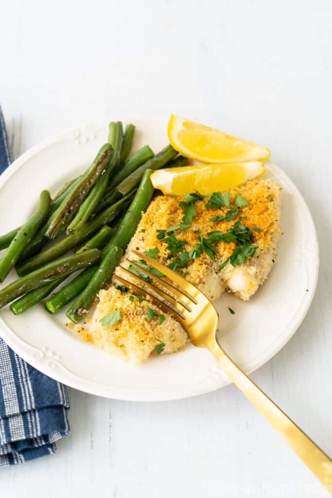 breaded flaky cod on a creamy plate with lemon slices and sauteed green beans