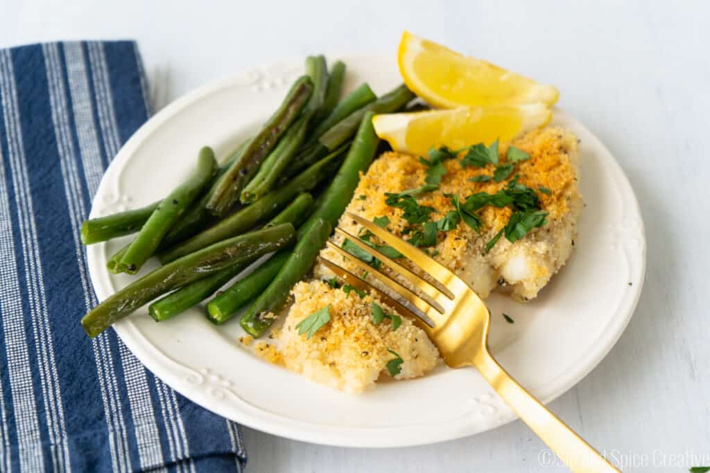 flaky white fish with breadcrumbs on a plate with green beans and lemon slices with a blue and white striped napkin.